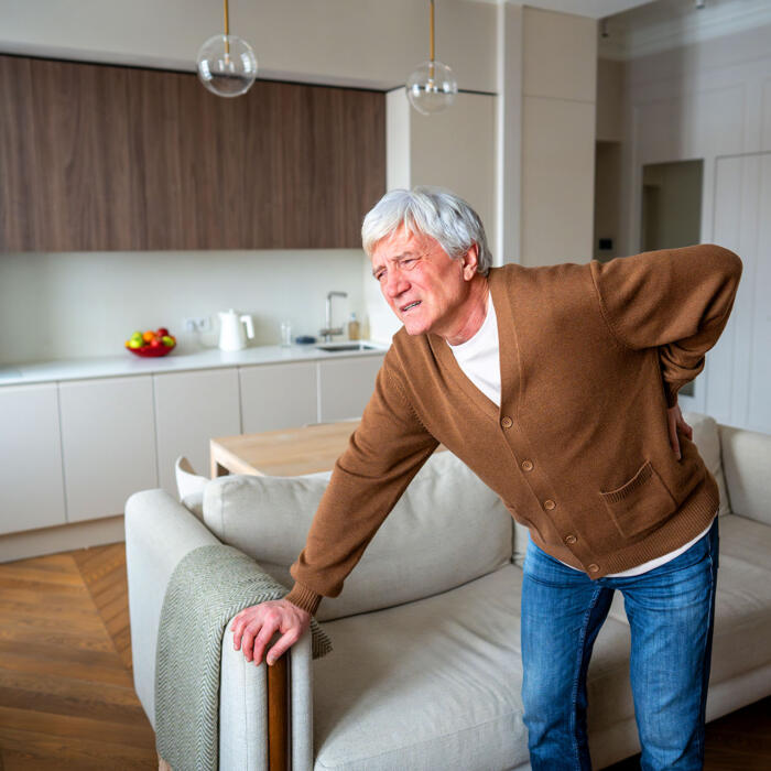 An older man standing in a living room while holding his lower back in pain.