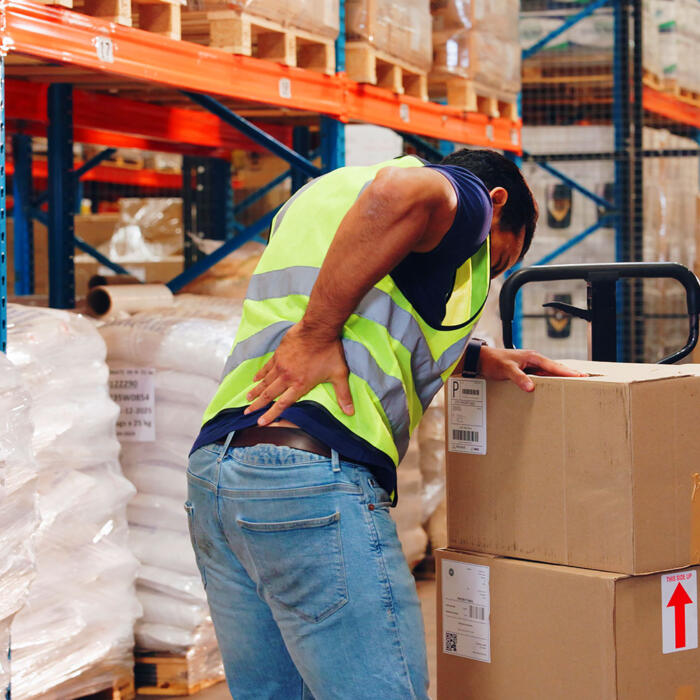 A warehouse worker in a high-visibility vest clutching his back while lifting heavy boxes.