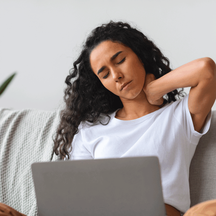 Young woman sitting on a sofa with a laptop and rubbing her neck due to muscle pain.